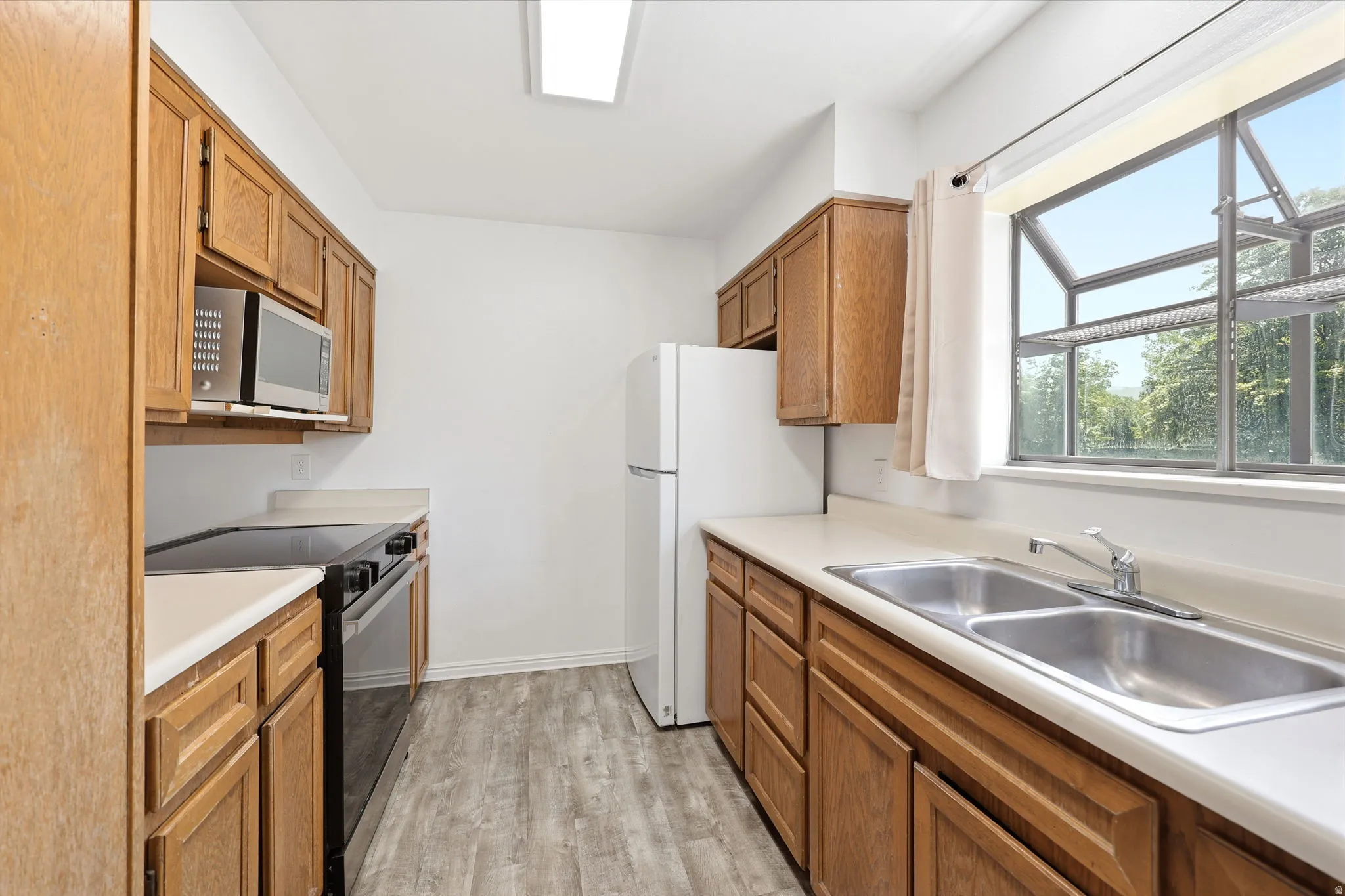 Kitchen featuring wood-finish cabinetry, light-toned countertops, a double-basin stainless steel sink, a stainless steel microwave, and wood-finish flooring
