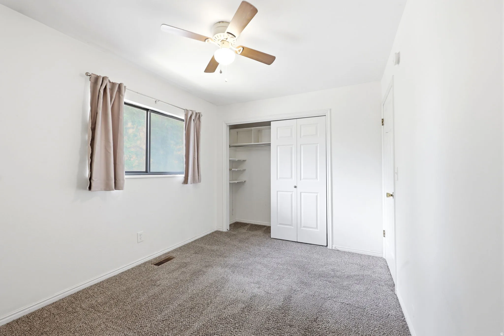 Carpeted interior room featuring a window with dark framing, a ceiling fan with wood-finish blades, a bi-fold closet door with shelving, and a white interior door