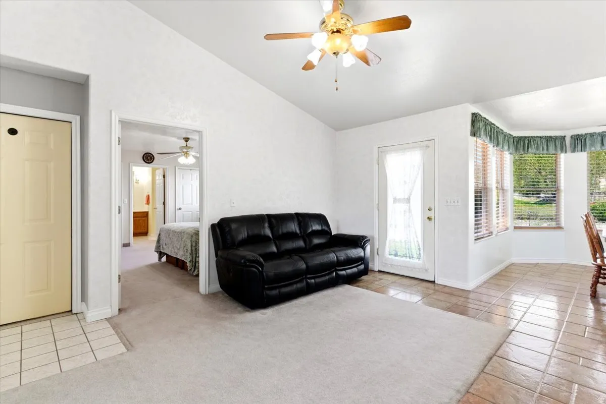 Living area featuring a textured white wall finish, a ceiling fan with light fixture, a bay window with horizontal blinds, and a combination of light-colored carpet and tiled flooring