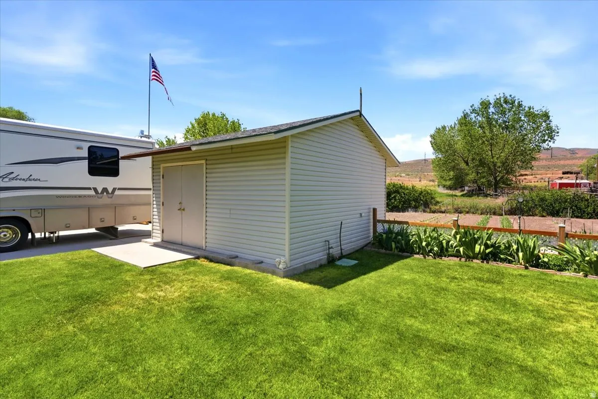Exterior storage shed with double doors and a concrete pad, situated on a grassy lawn with a wood fence bordering a garden area
