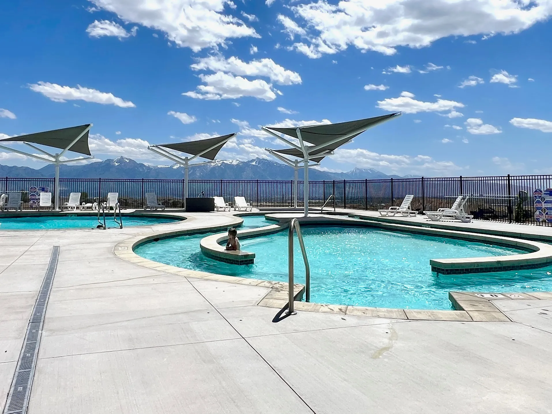 Expansive outdoor swimming pool area with multiple water features, sun loungers, and shade structures