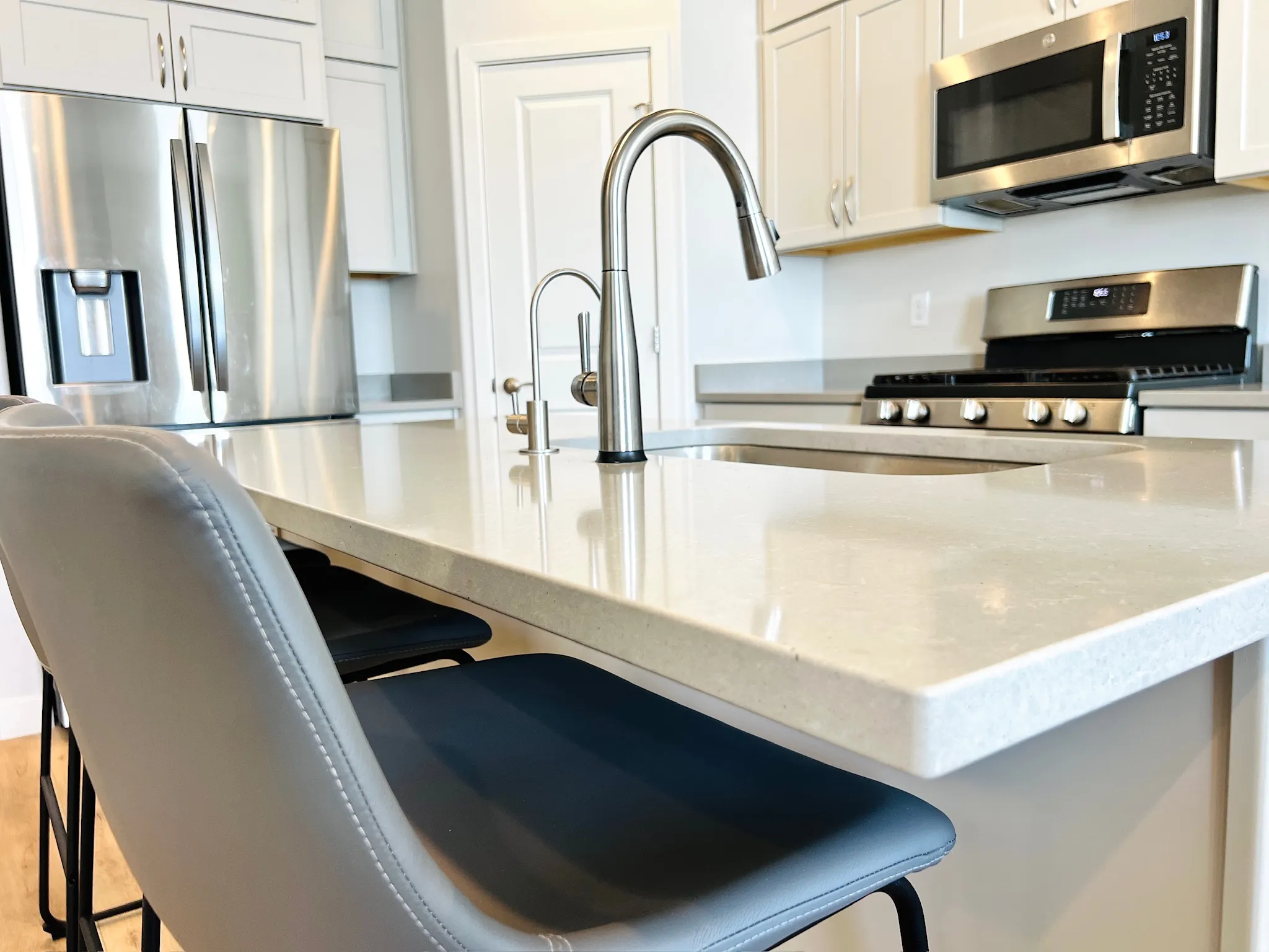 Kitchen island featuring an undermount sink with a gooseneck faucet, light-toned countertops, white cabinetry, and stainless steel appliances including a refrigerator, range, and microwave