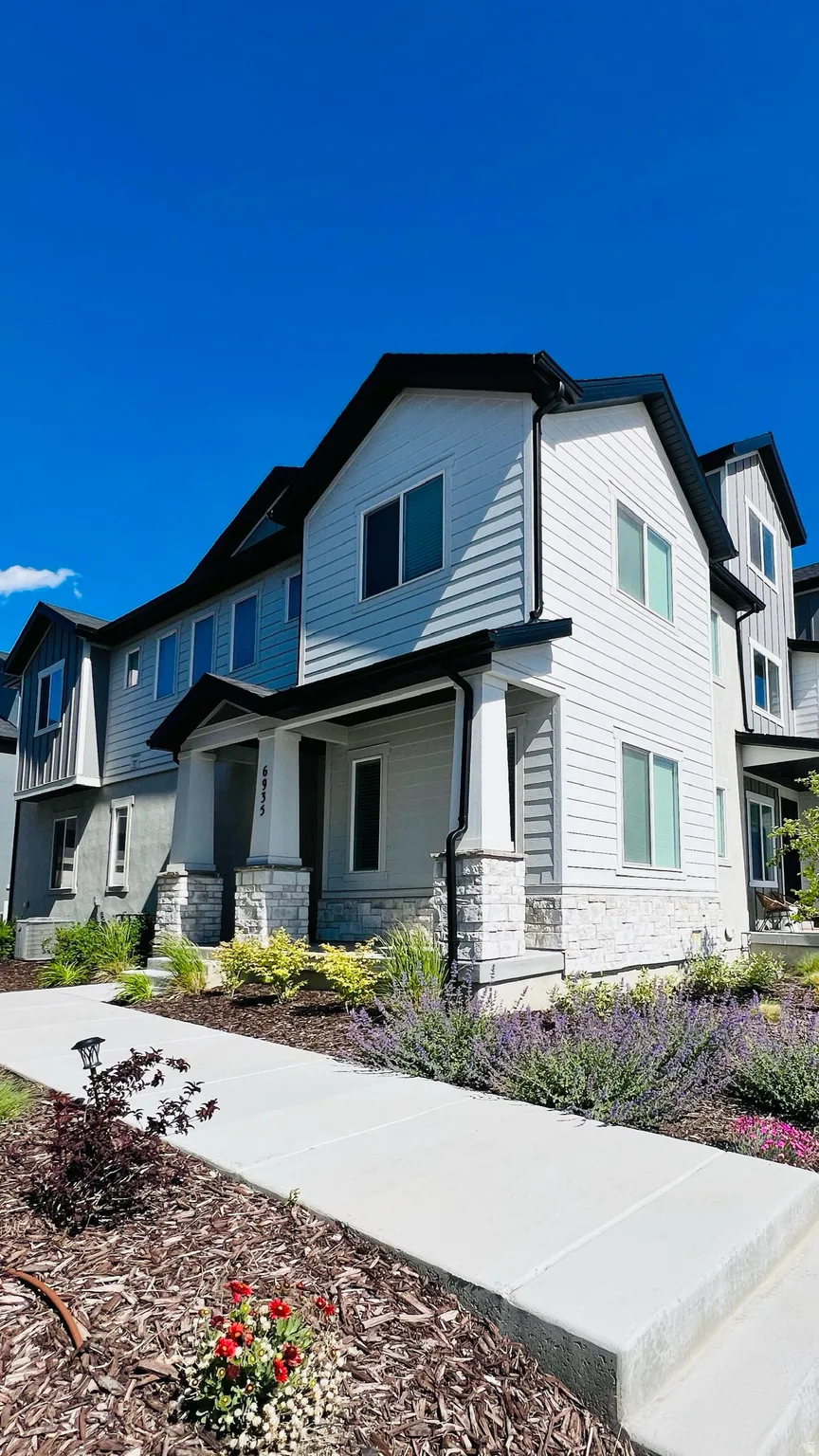 Modern townhome exterior featuring light-colored siding with dark trim, a covered porch with stone-clad columns, a clear concrete walkway, and landscaped garden beds with mulch and plantings