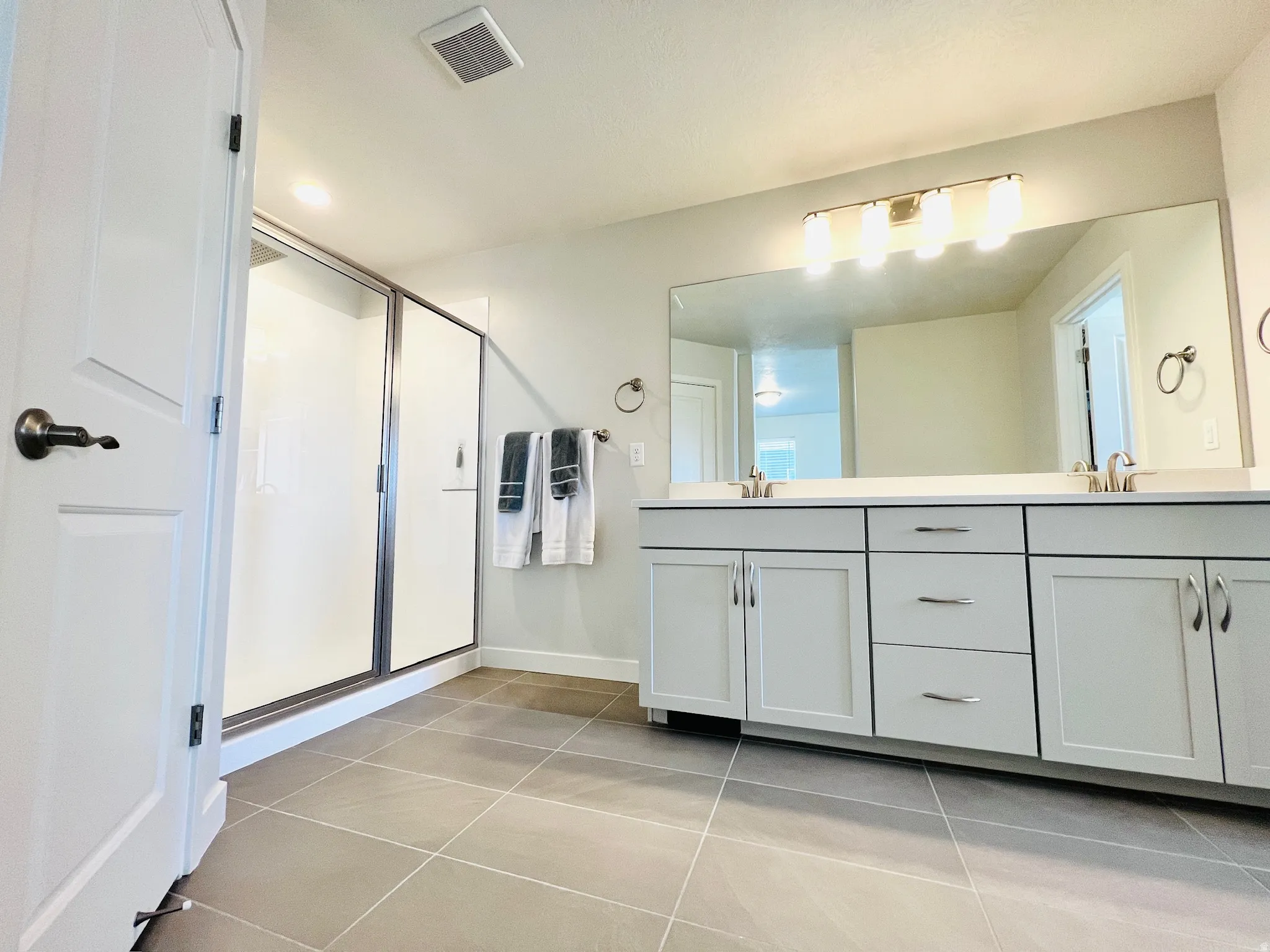 Spacious bathroom featuring a dual vanity with white cabinetry and chrome hardware, a wide mirror, a glass-enclosed shower, and large format floor tiling