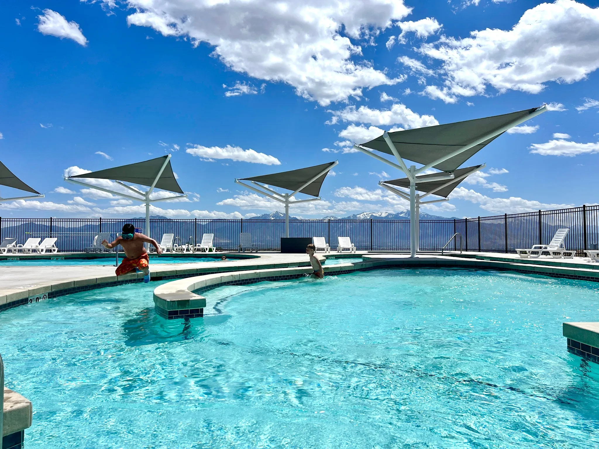 Resort-style pool area featuring multiple shade structures, integrated lounging ledges, and a perimeter fence