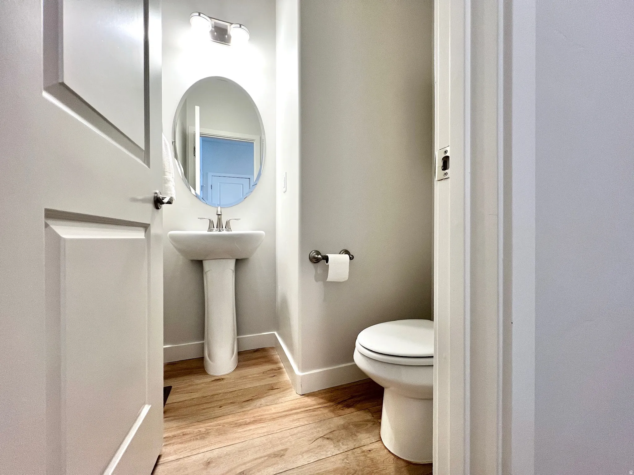 Powder room featuring a pedestal sink with chrome faucet, an oval mirror, and wood-finish flooring