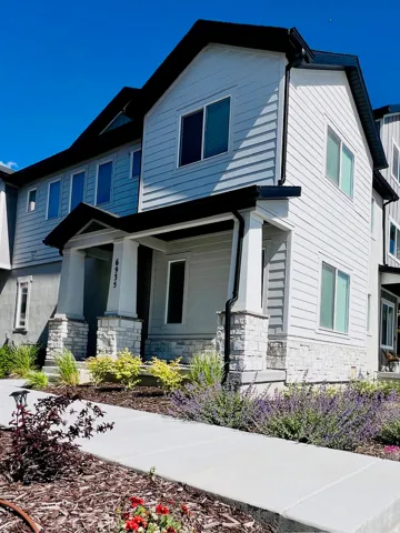 Modern multi-level residence featuring light-colored siding, dark trim accents, stone veneer detailing on the porch columns and base, a concrete walkway, and landscaped garden beds with various plantings