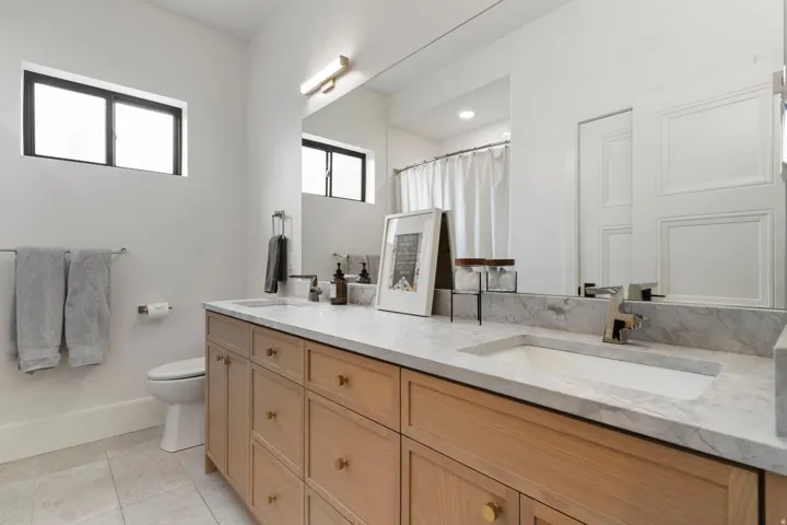 Double vanity featuring light wood cabinetry with gold-tone hardware, a light stone countertop, and two integrated rectangular sinks with modern chrome faucets