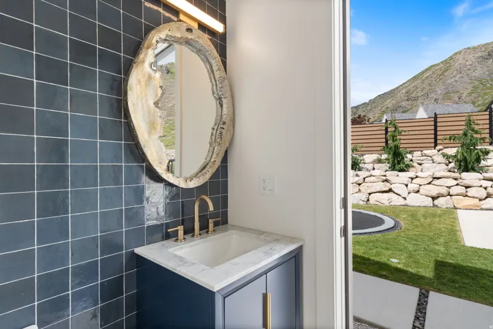 Pool bathroom vanity with a white countertop and gold-toned faucet, featuring a unique mirror and dark tiled wall