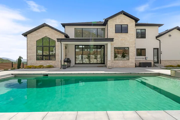 Contemporary residence featuring a rectangular swimming pool, expansive concrete patio, light-toned stone and stucco exterior, large black-framed windows, and a covered patio area