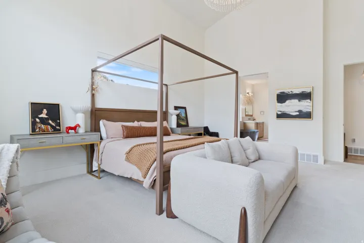 Vaulted ceilings highlight this expansive bedroom featuring a four-poster bed frame, neutral-toned carpeting, and high windows providing natural illumination
