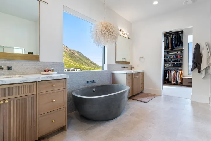 Freestanding soaking tub with a wall-mounted faucet, dual wood-finish vanities with marble countertops, large picture window with mountain scenery, and a custom built-in closet system