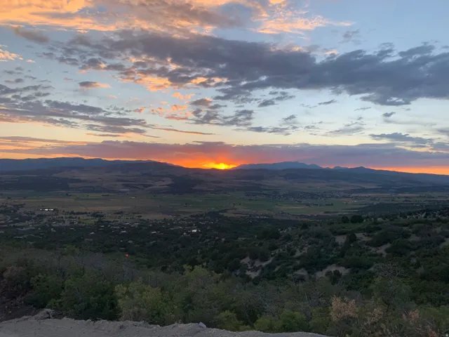 Expansive mountain and valley landscape at sunset, featuring a striking orange sun on the horizon, varied terrain with scattered trees, and distant structures