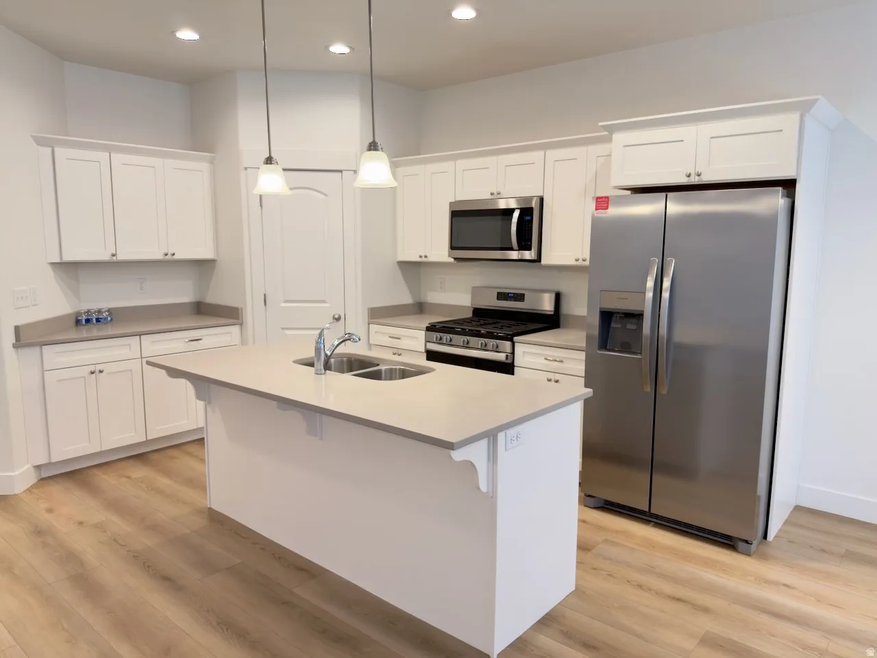 Kitchen featuring light wood-finish flooring, white cabinetry, a central island with a double basin sink, stainless steel appliances, and pendant lighting