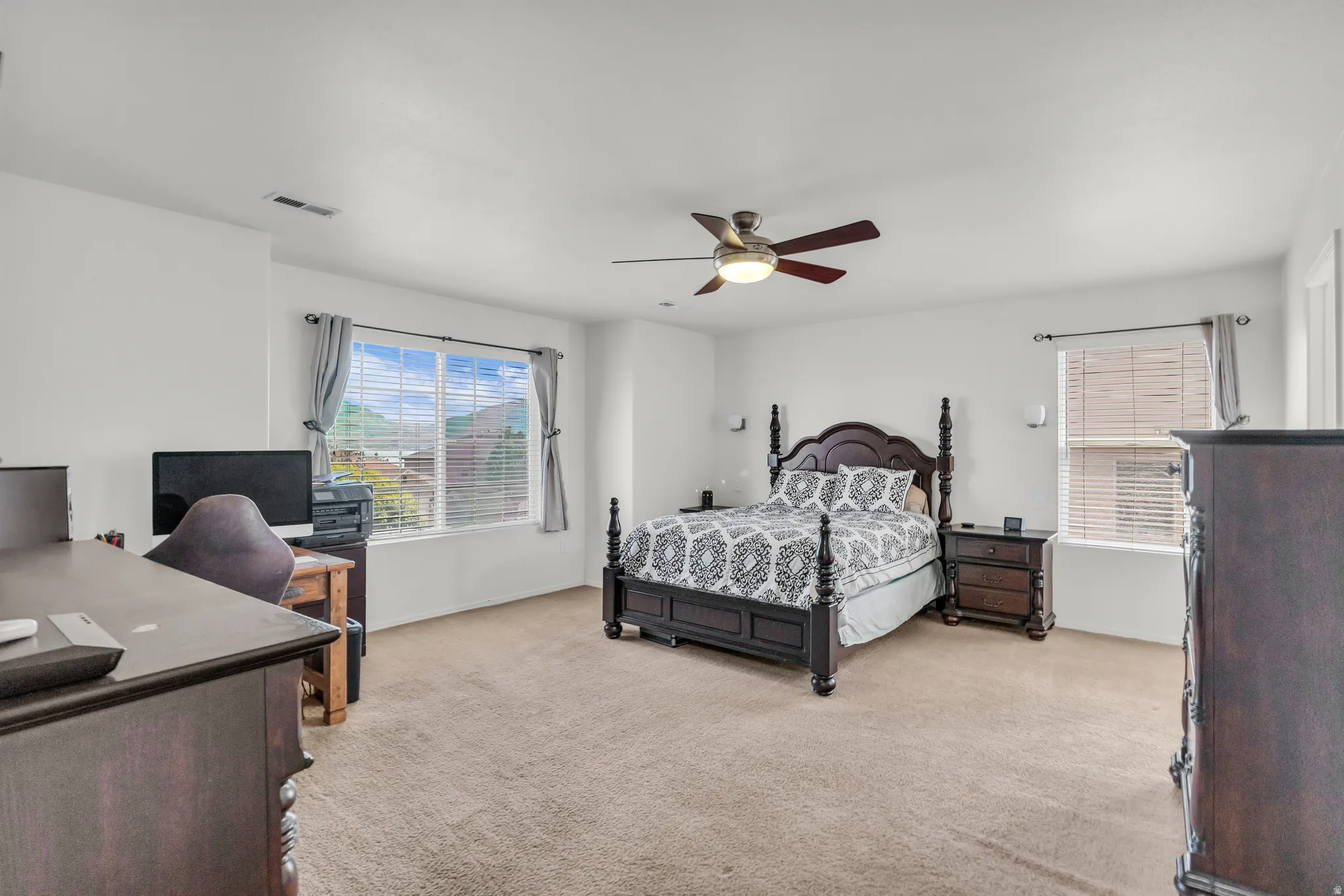Spacious room featuring neutral carpet flooring, white walls, two windows with blinds, a ceiling fan with integrated lighting, and a built-in desk area