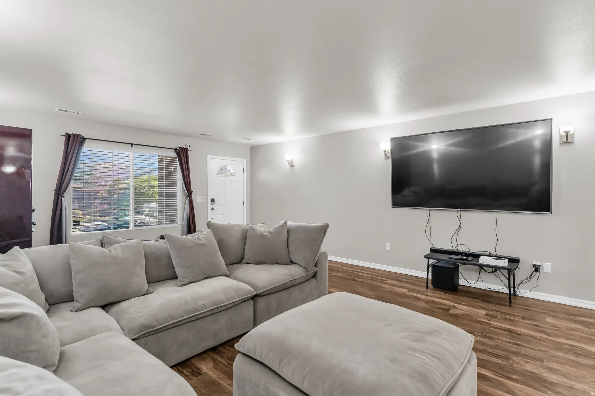 Living area featuring wood-finish flooring, a white paneled entry door, and a large window with blinds