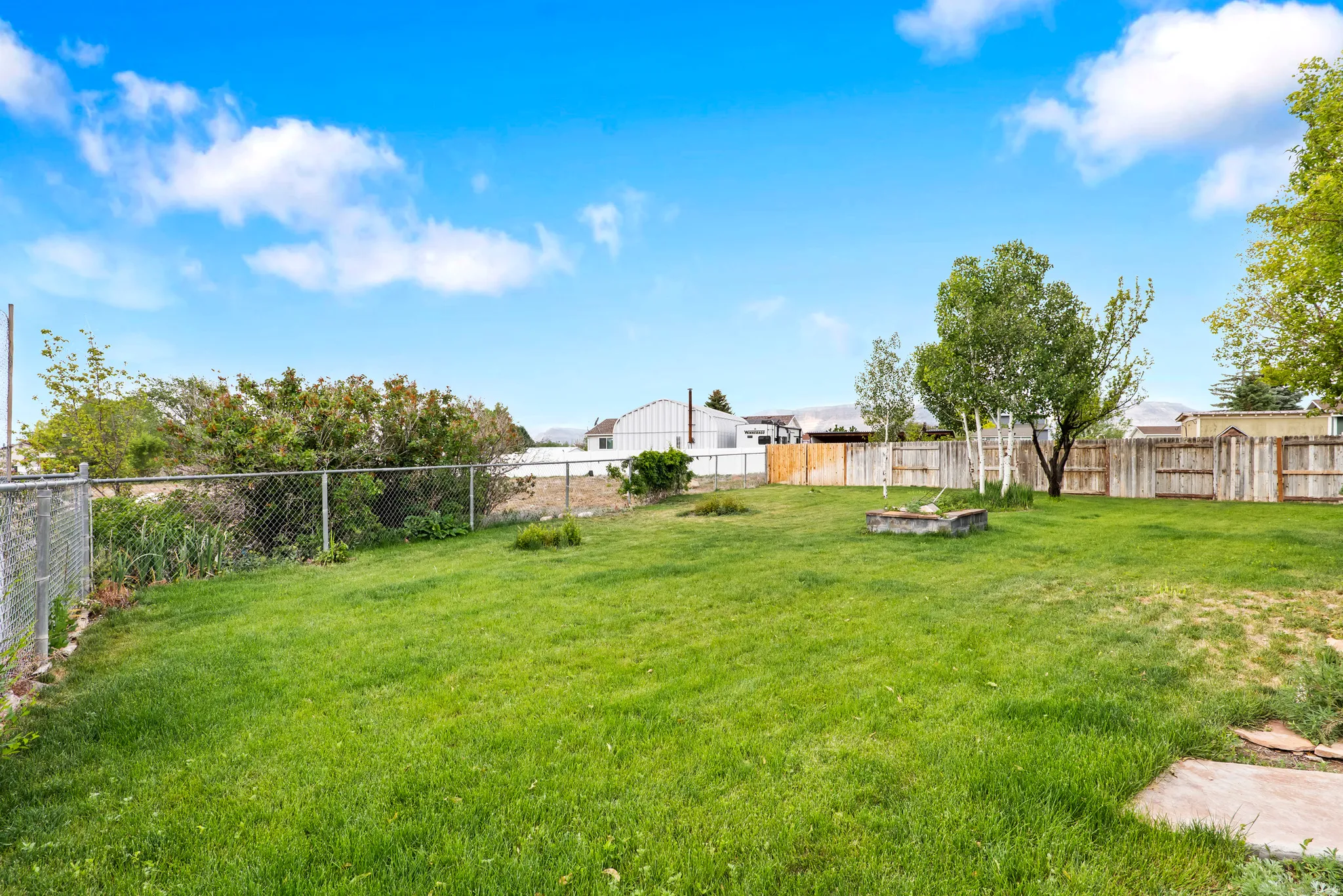 Expansive grassy yard featuring a chain-link fence, wood privacy fencing, mature trees, and a built-in fire pit