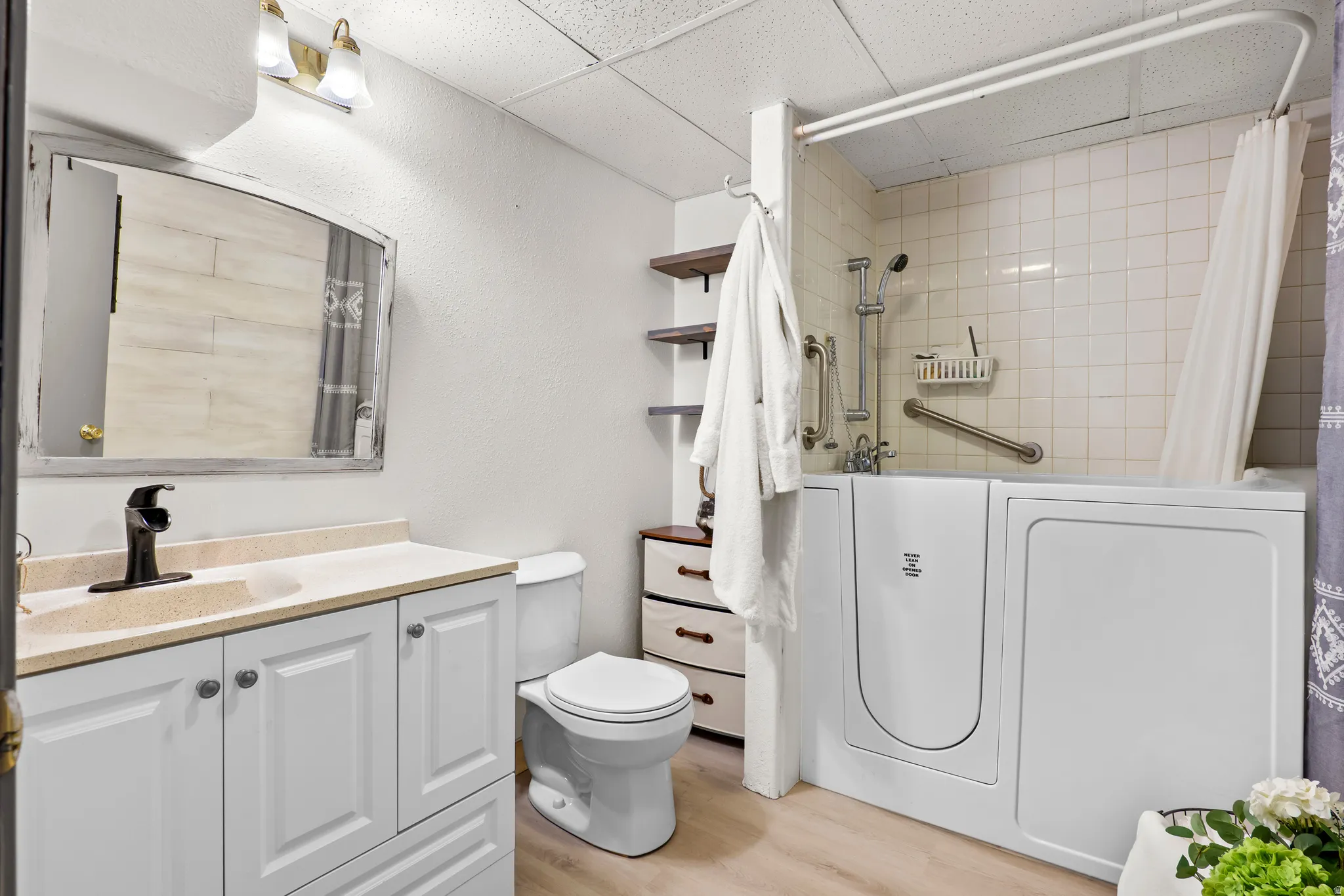 Bathroom featuring a walk-in tub with grab bars, white tiled shower surround, a vanity with a light-colored countertop and white cabinetry, a toilet, and light wood-finish flooring
