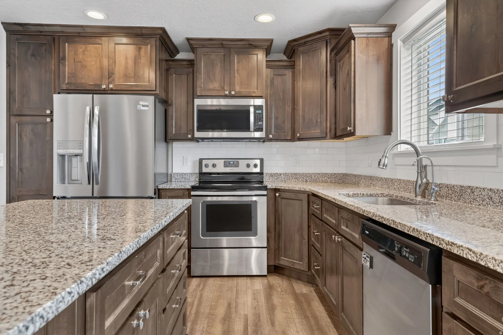 Kitchen featuring granite countertops, wood-finish flooring, dark wood cabinetry, a stainless steel appliance suite, and a white subway tile backsplash