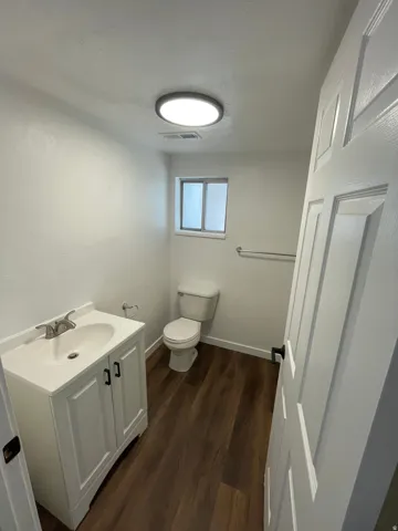 Bathroom featuring wood-finish flooring, a white vanity with paneling and dark hardware, a white toilet, and a window with obscured glass