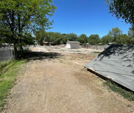 Expansive outdoor area featuring a gravel and dirt surface, a chain-link fence, mature trees, and a freestanding shed with a white exterior and green shingle roof