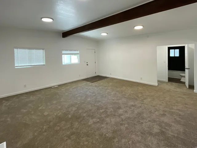 Spacious room featuring neutral carpeting, white painted walls, and exposed ceiling beams