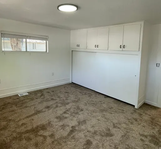 Room featuring neutral-toned carpeting, a window with blinds, and a built-in wall unit with overhead cabinetry and a lower panel