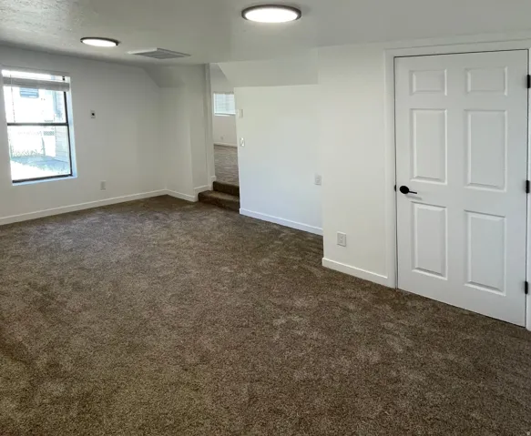 Spacious carpeted room featuring a six-panel interior door with matte black hardware, recessed wall outlets, and two flush-mount ceiling lights