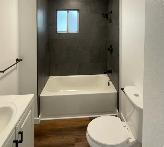 Bathroom featuring a white bathtub with dark tiled surround, a frosted window, matte black shower fixtures, a white vanity with integrated sink, and wood-finish flooring