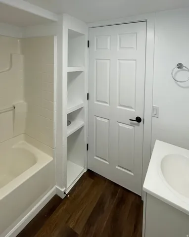 Bathroom featuring a tub/shower combination with tile surround, built-in shelving, a white paneled door with black hardware, and wood-finish flooring