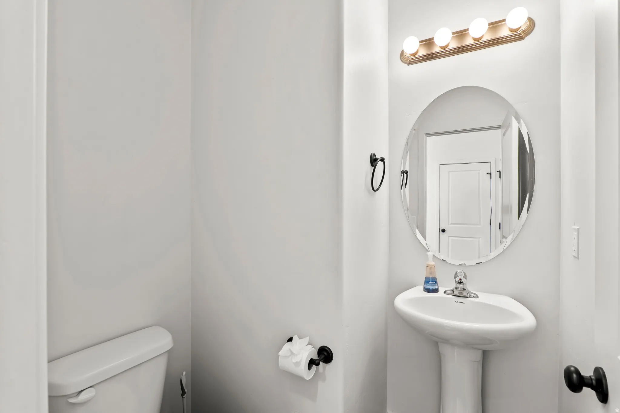 Pedestal sink with chrome faucet, complemented by a round framed mirror and a four-bulb vanity light fixture