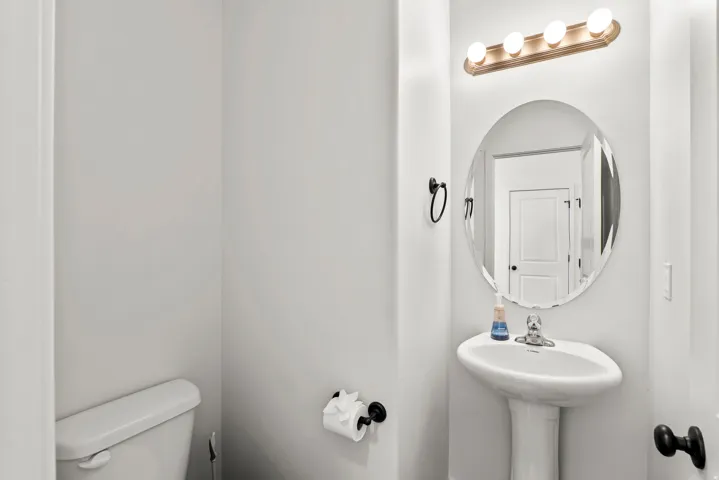 Pedestal sink with chrome faucet, complemented by a round framed mirror and a four-bulb vanity light fixture