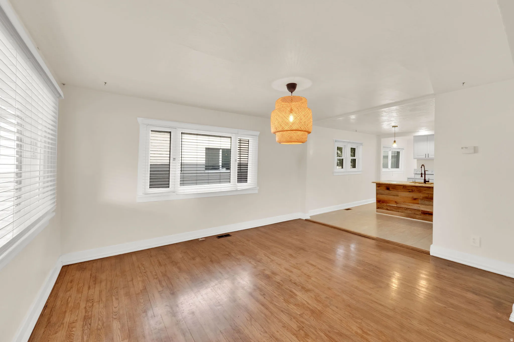 Spacious room featuring wood-finish flooring, multiple windows with blinds, a woven pendant light fixture, and white painted walls
