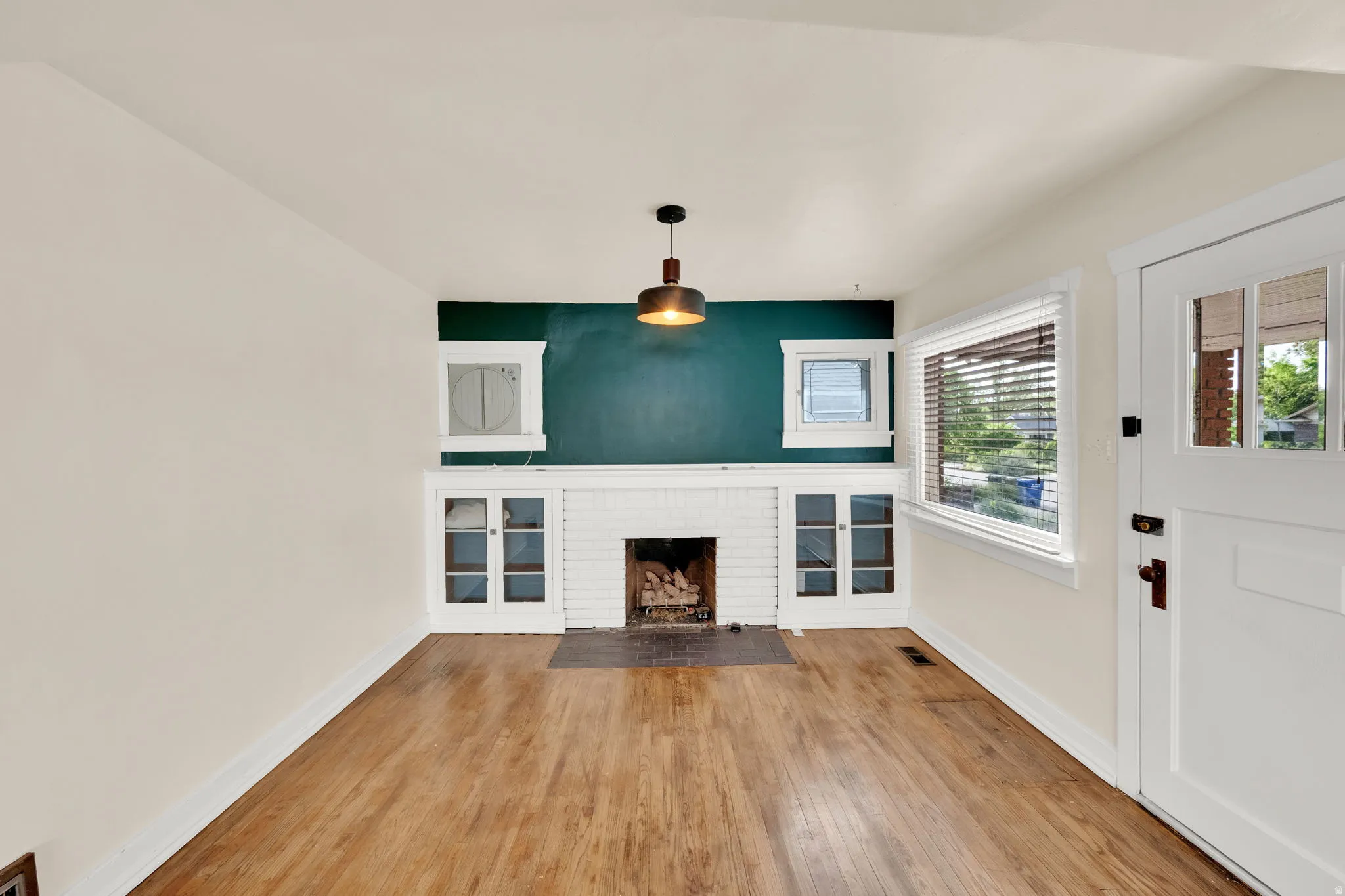 Living space featuring wood-finish flooring, a white brick gas fireplace with a dark hearth, built-in cabinetry with glass panel doors, and a decorative pendant light fixture