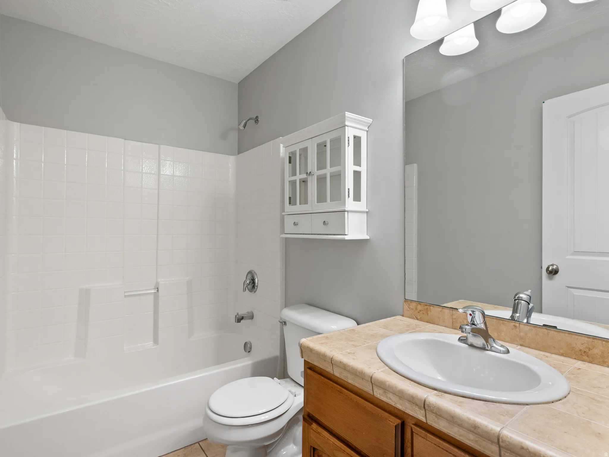 Bathroom featuring a white bathtub/shower combination with tiled surround, a white toilet, and a wood-finish vanity with a tiled countertop and integrated sink