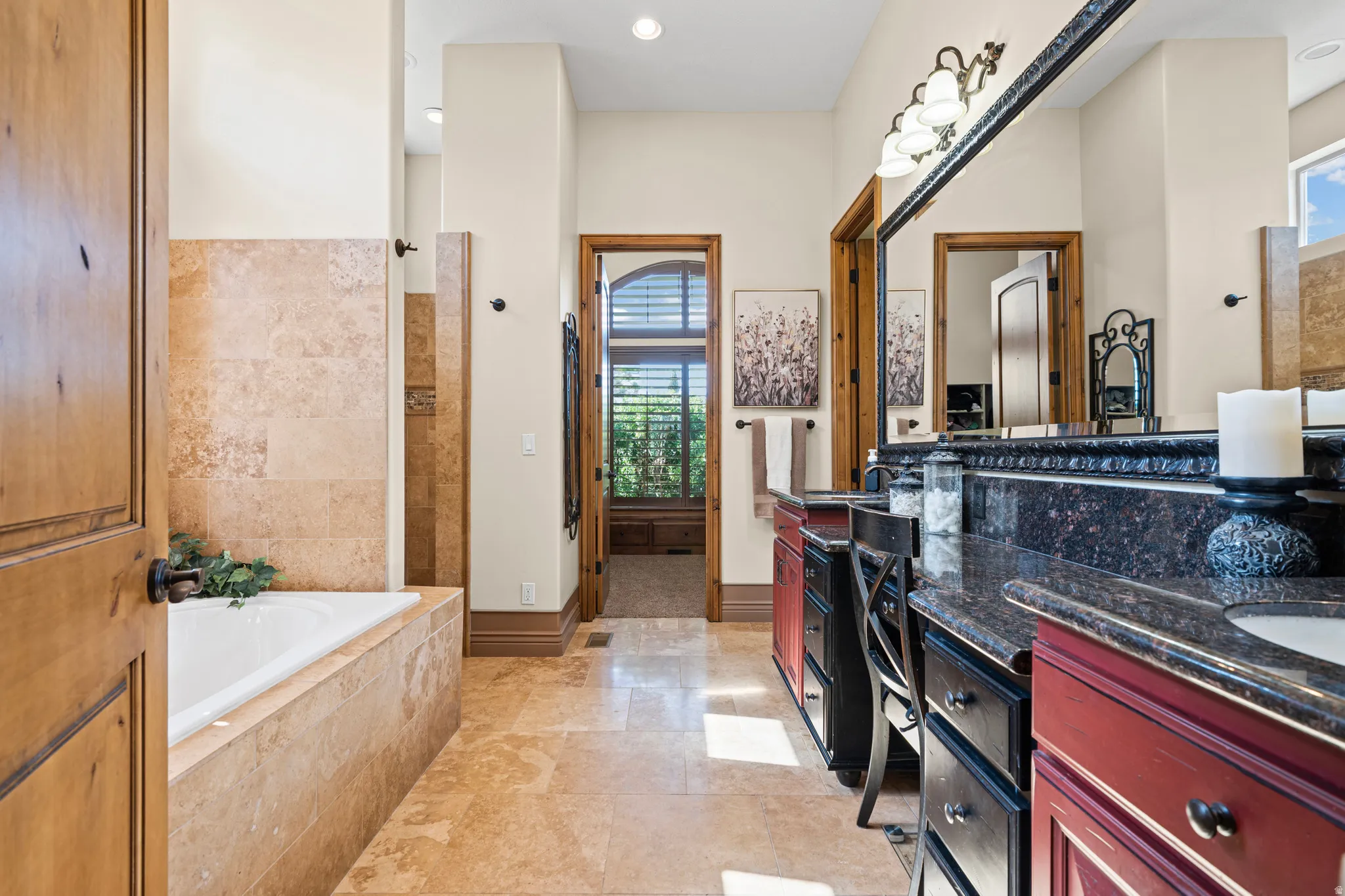 Spacious bathroom featuring a built-in tub with natural stone tile surround, natural stone tile flooring, an oversized vanity with dual sinks, dark countertops, and wood-finish cabinetry