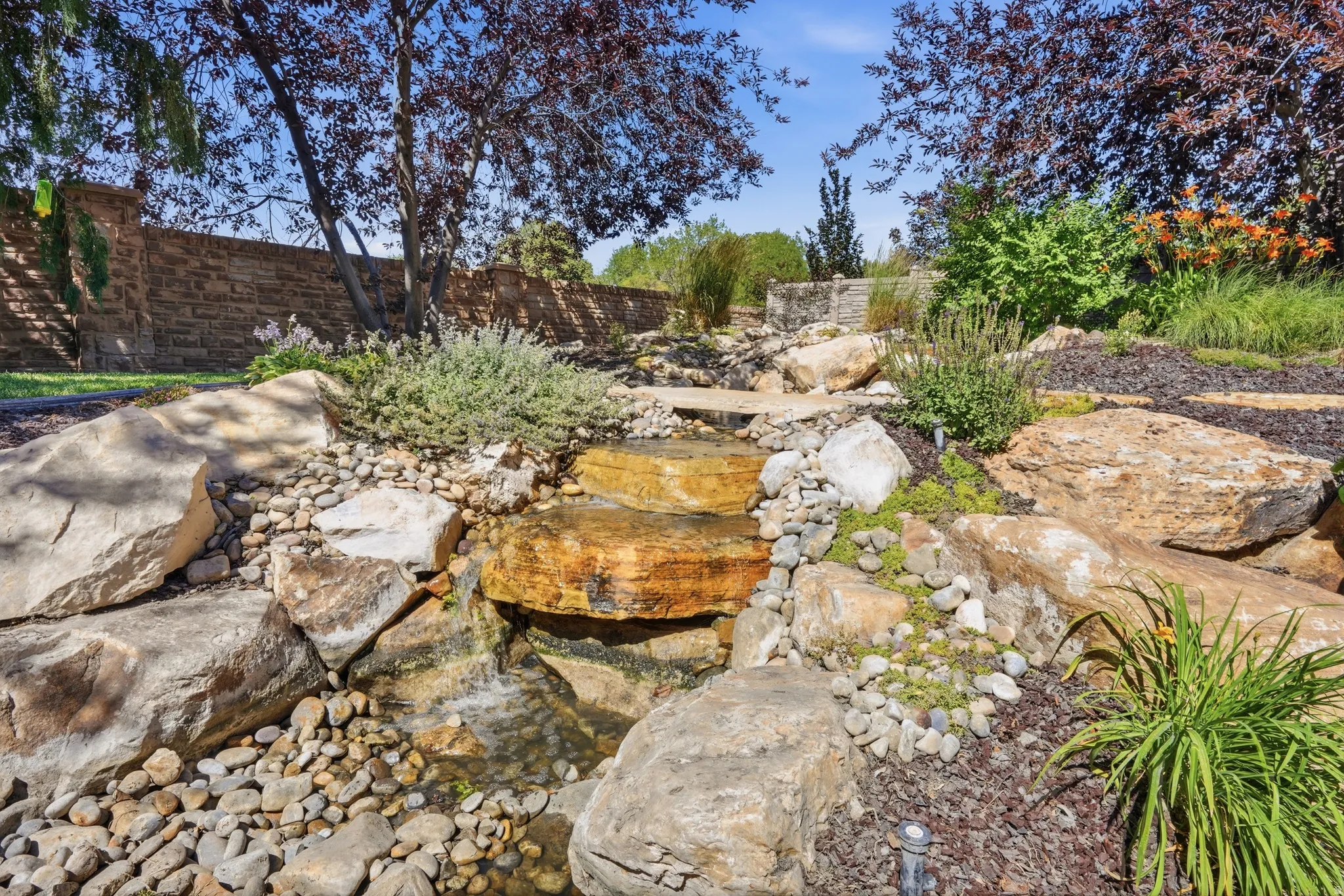 Landscaped waterfall with natural rock formations and tiered waterfalls, surrounded by various plantings, gravel, and bark mulch