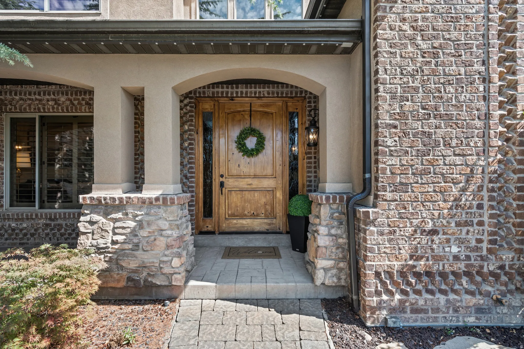 Arched entryway featuring a substantial wood door with sidelights, brick and stone facade, paver walkway, and exterior lighting fixture