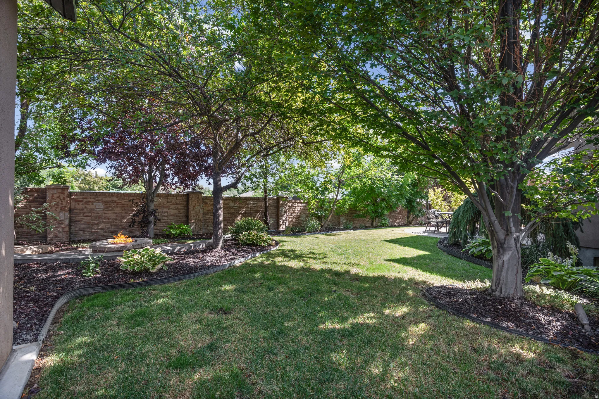 Expansive backyard featuring a manicured lawn, mature trees, a stone fire pit, and a brick privacy fence