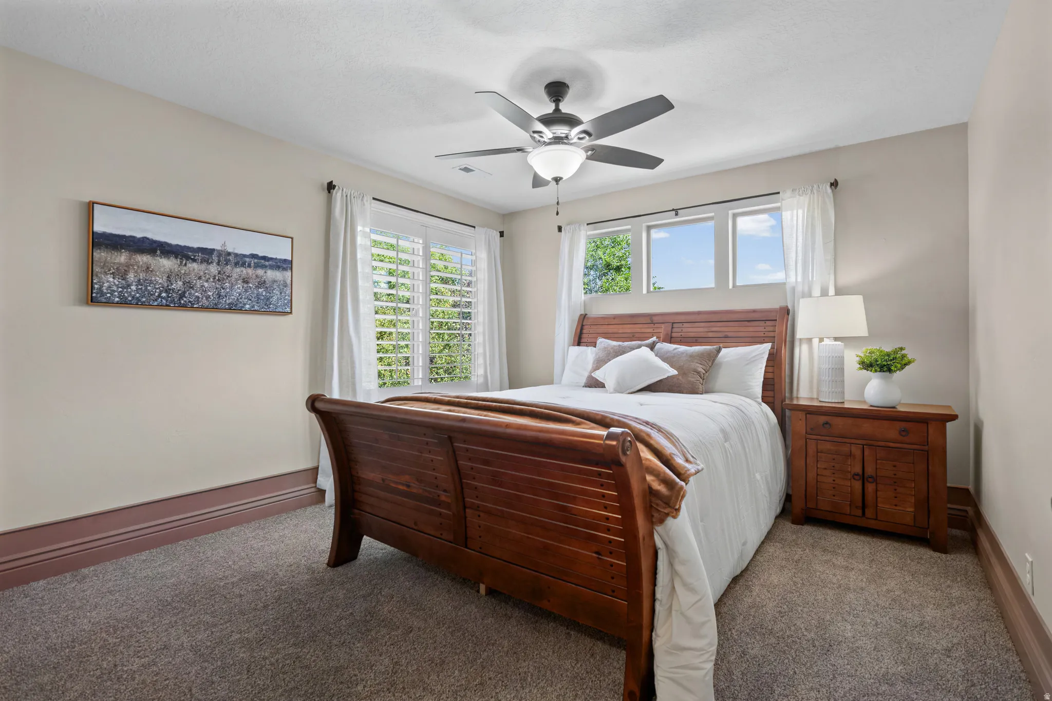 Bedroom featuring a dark wood sleigh bed, a ceiling fan with integrated lighting, and multiple windows with plantation shutters and white curtains