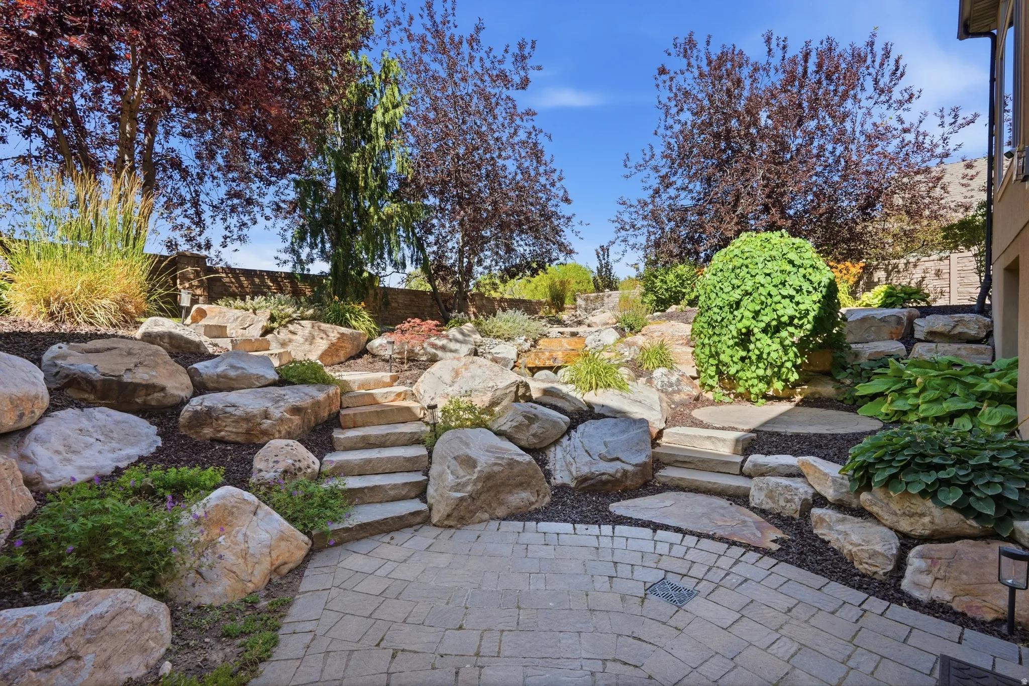 Paver patio featuring a terraced rock garden with stone steps, mature trees, and diverse plantings