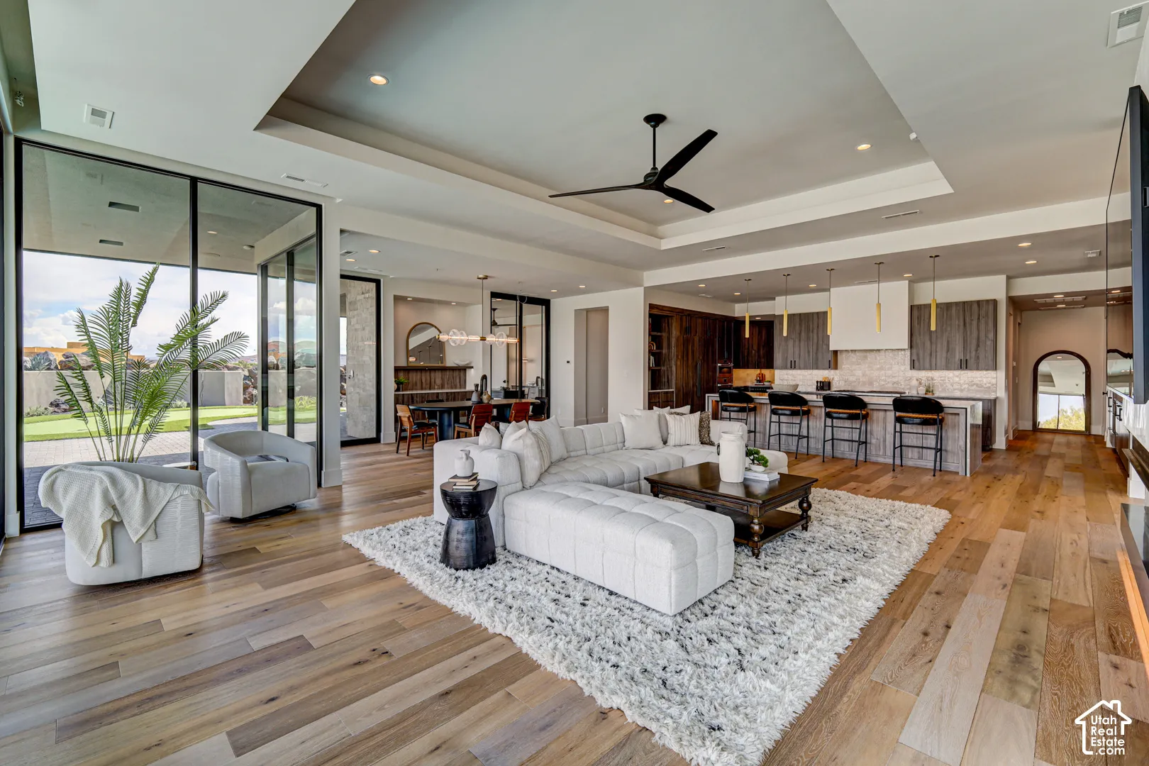Expansive wood-finish flooring throughout, tray ceiling with recessed lighting and ceiling fan, full-height glass patio doors, and a kitchen featuring a large island with bar seating and pendant lighting