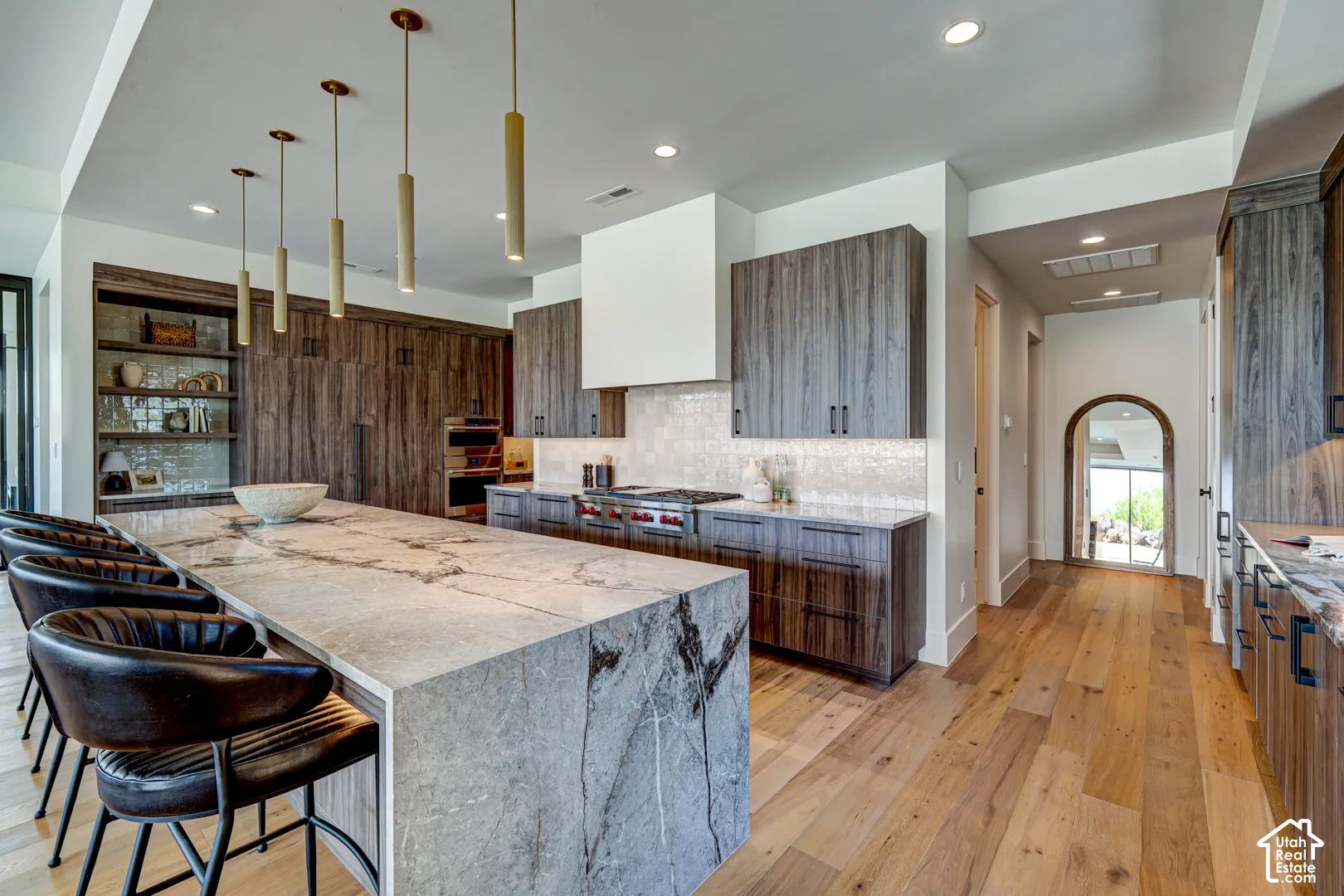 Large central island with stone countertop, wood-finish flooring, custom wood cabinetry, recessed lighting, and modern pendant fixtures