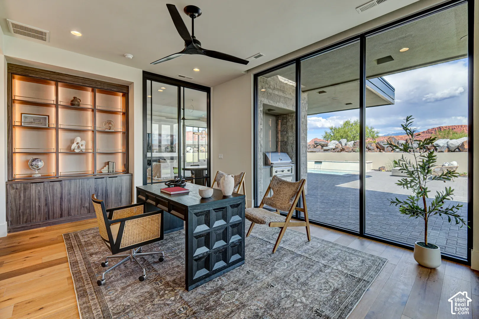 Wood-finish flooring, built-in shelving with integrated lighting, a black ceiling fan, floor-to-ceiling glass doors, and an outdoor kitchen with a built-in grill