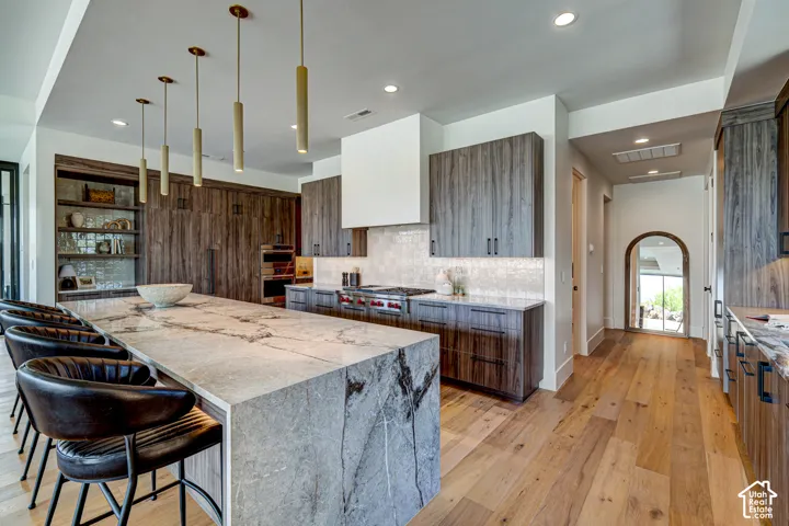 Large central island with stone countertop, wood-finish flooring, custom wood cabinetry, recessed lighting, and modern pendant fixtures