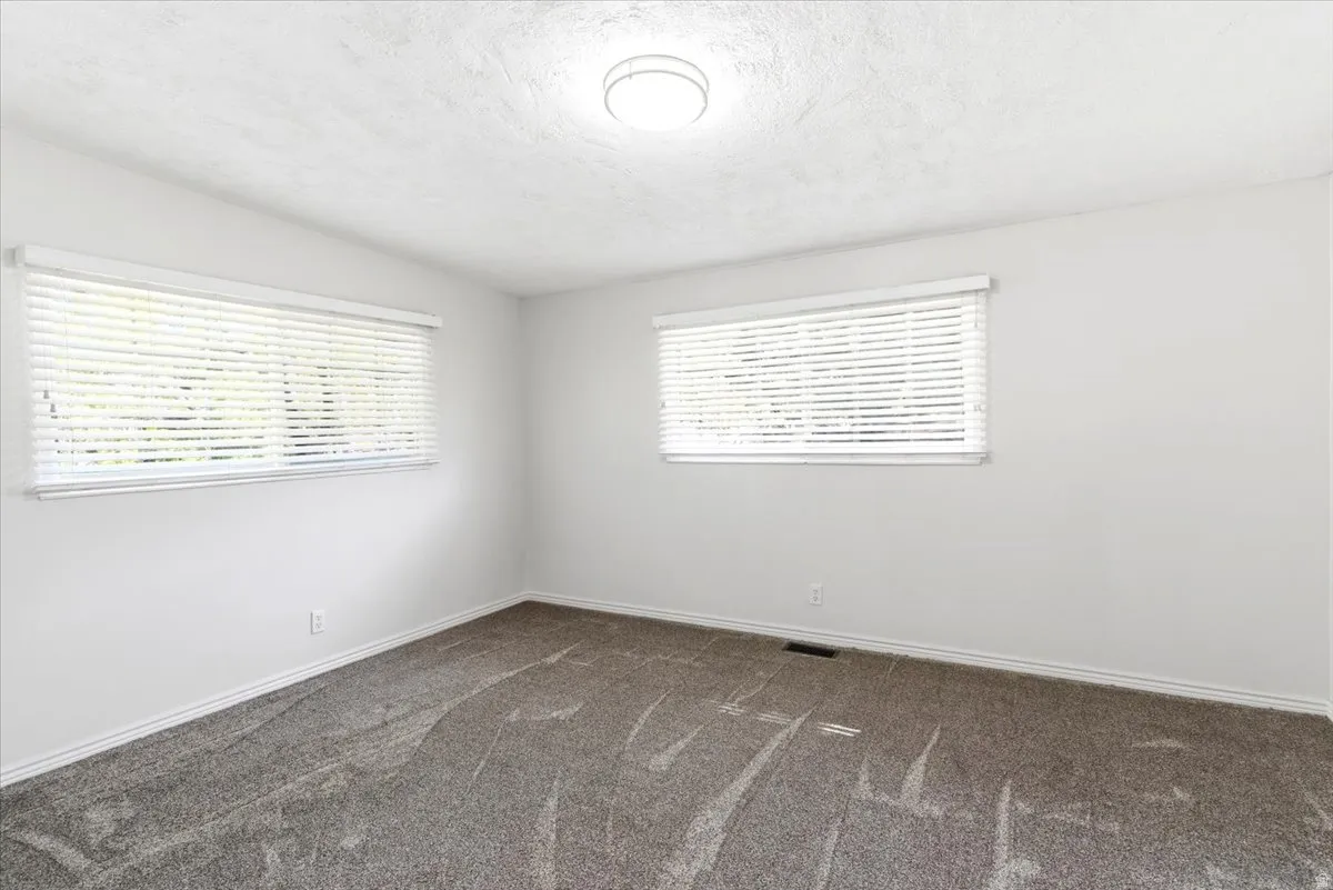 Bright room with white walls, two windows featuring horizontal blinds, and a neutral-toned carpeted floor
