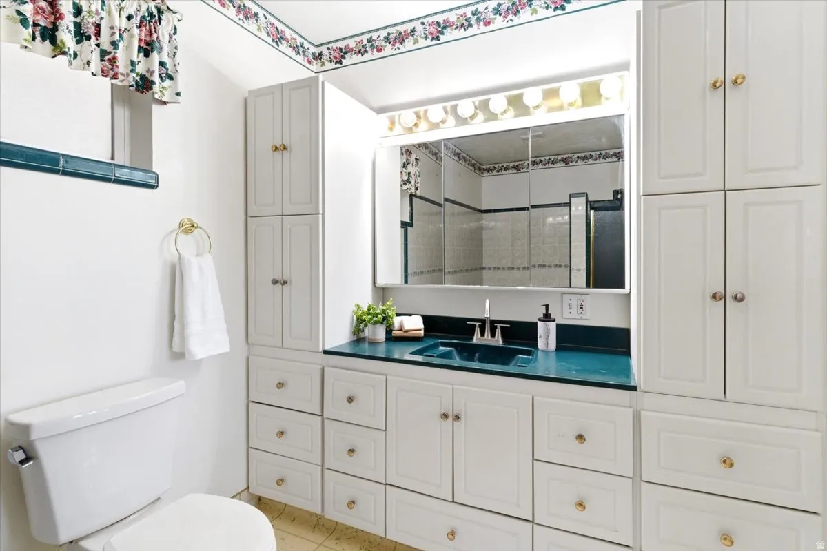 Bathroom featuring an extensive white cabinetry system, a teal countertop with an integrated sink, a large vanity mirror with overhead lighting, and brass-tone hardware