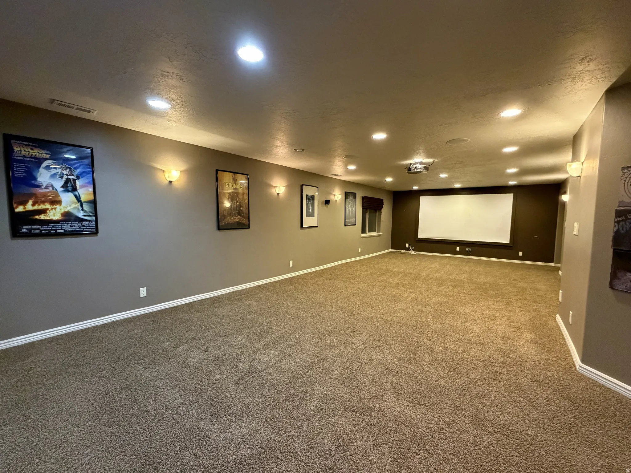 Spacious room featuring extensive recessed lighting, sconce lighting, an integrated projector, a large projection screen, and textured carpeting