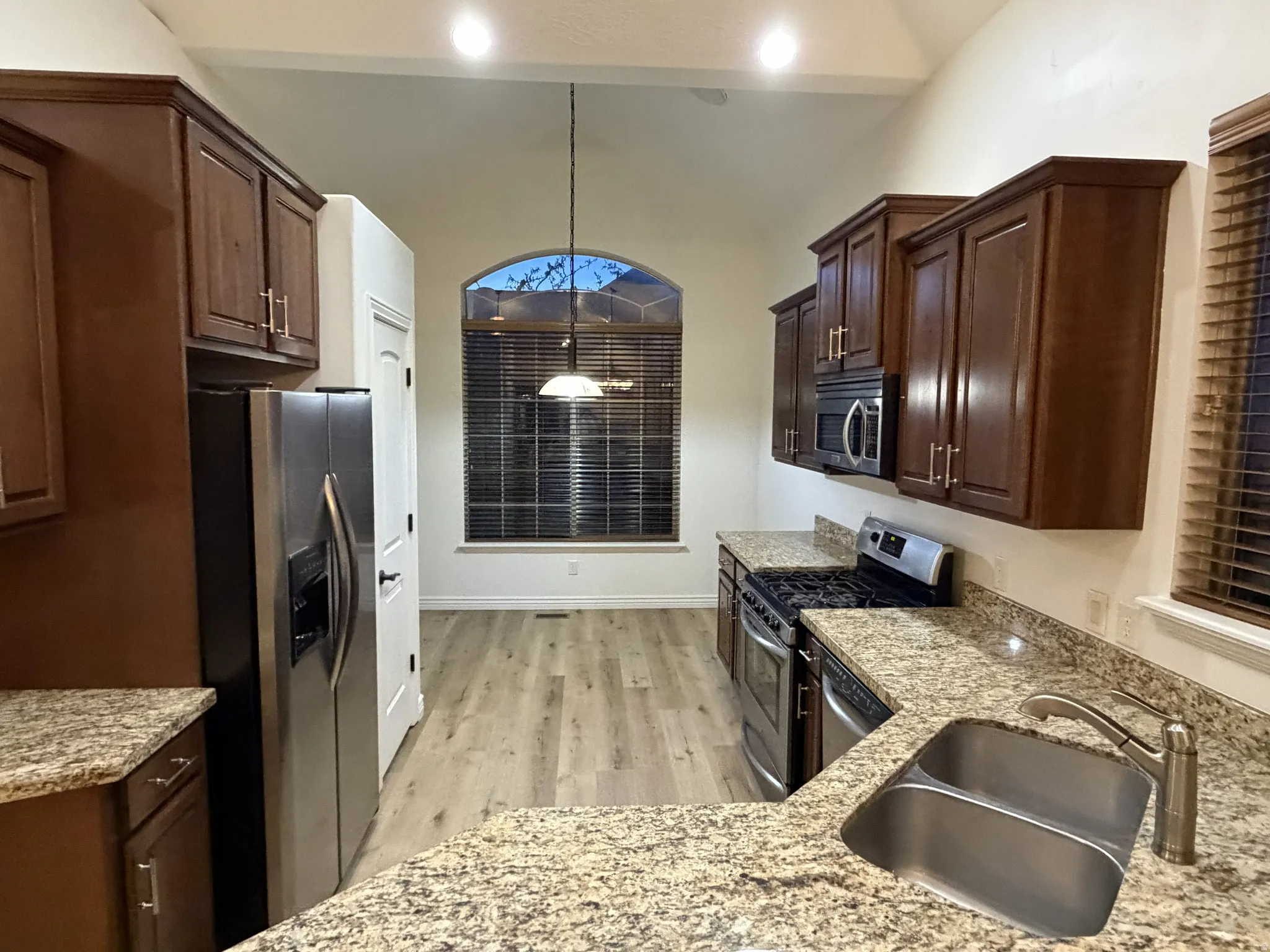Kitchen featuring granite countertops, wood-finish flooring, stainless steel appliances, dark wood cabinetry, and an arched window with blinds
