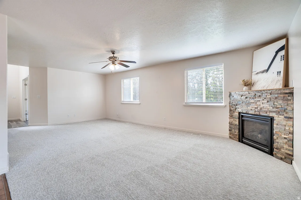 Spacious living area featuring light-colored carpeting, a stone-clad fireplace, two windows with blinds, a ceiling fan with integrated lighting, and white walls